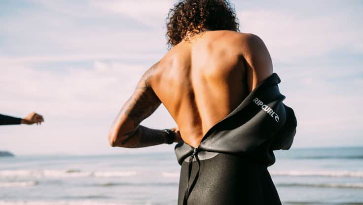 Frau geht am Strand entlang in ihrem Neoprenanzug und dem Surfbrett unterm Arm